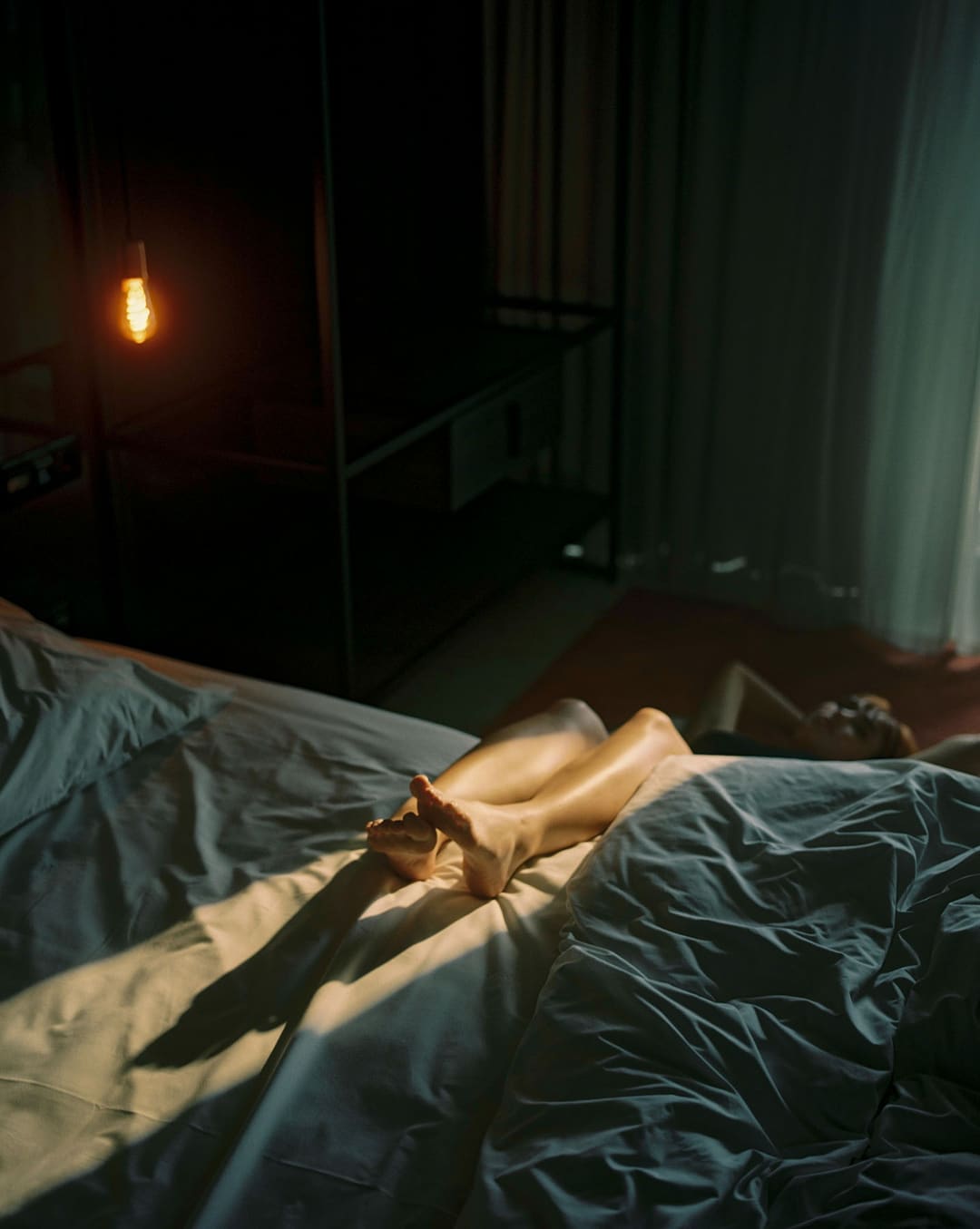 Um quarto escuro com uma lâmpada acesa ao fundo: numa cama de lençóis azul-acinzentados, um raio de sol ilumina as pernas cruzadas de uma mulher deitada.