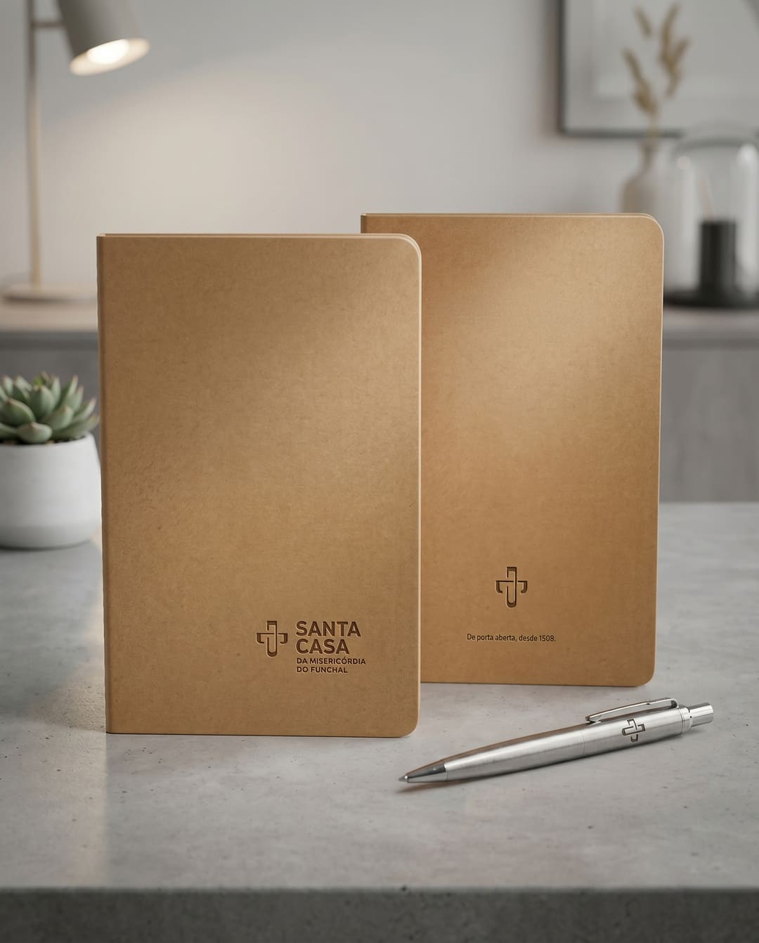 Dois cadernos de capa kraft com a marca gravada, «De porta aberta, desde 1508.», e uma caneta com o símbolo.