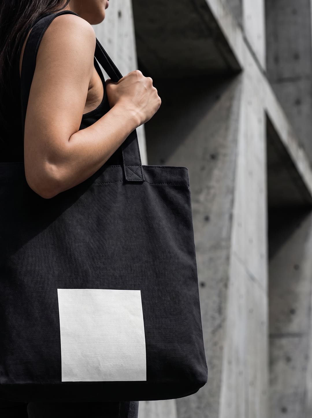 Uma tote bag preta ao ombro, com um rectângulo branco em baixo, diante de uma parede de betão.