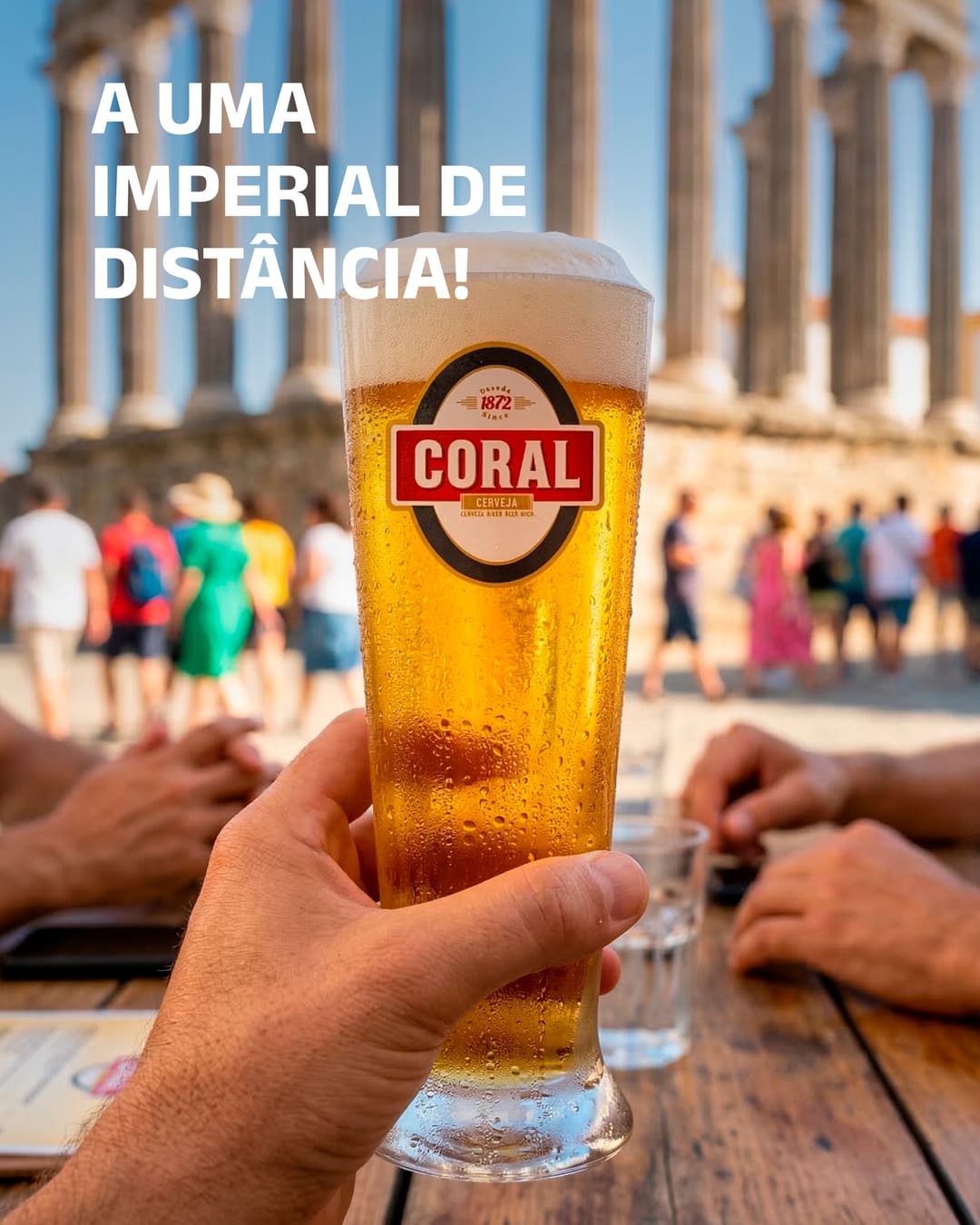 Uma mão levanta uma imperial Coral diante das colunas do Templo Romano de Évora, com visitantes ao fundo. Em cima, «A uma imperial de distância!».