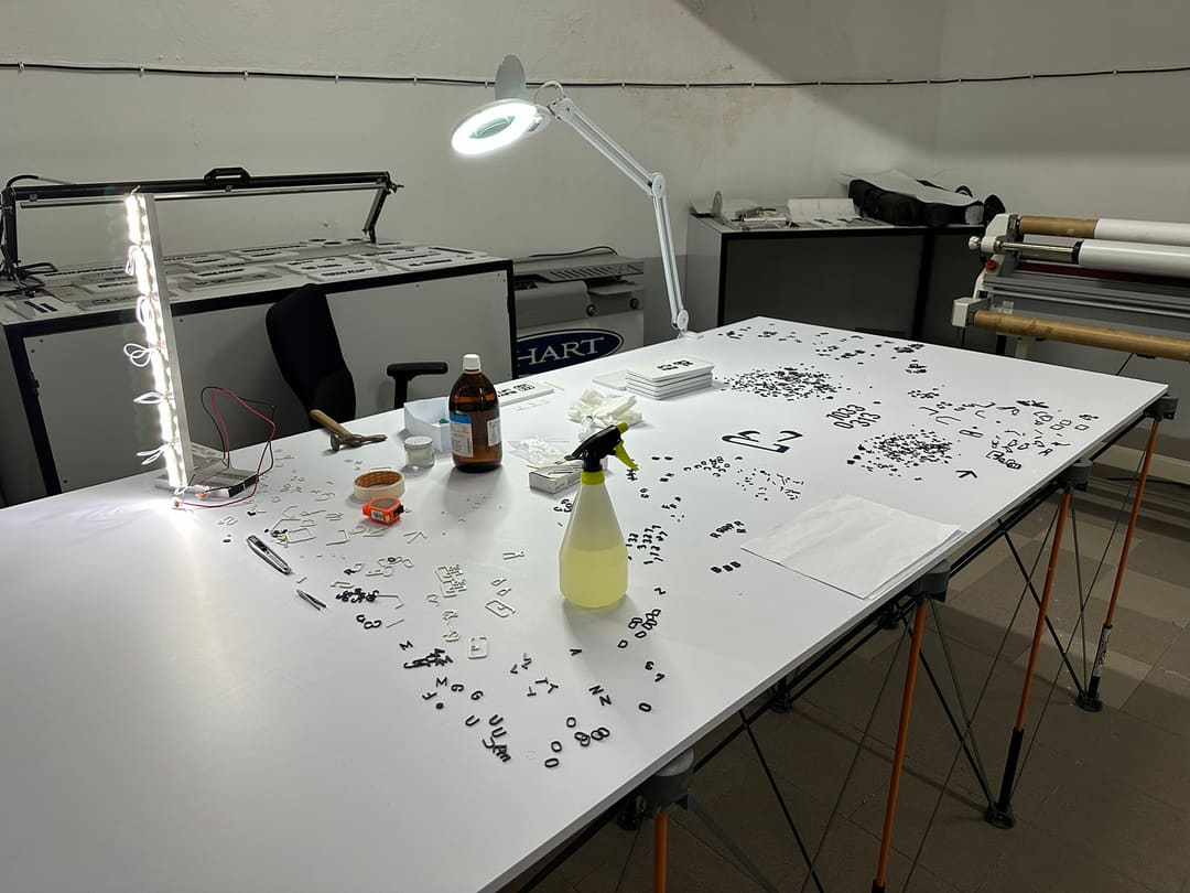 A mesa da oficina com a lupa de bancada, letras e números espalhados, placas empilhadas e uma barra de LED.