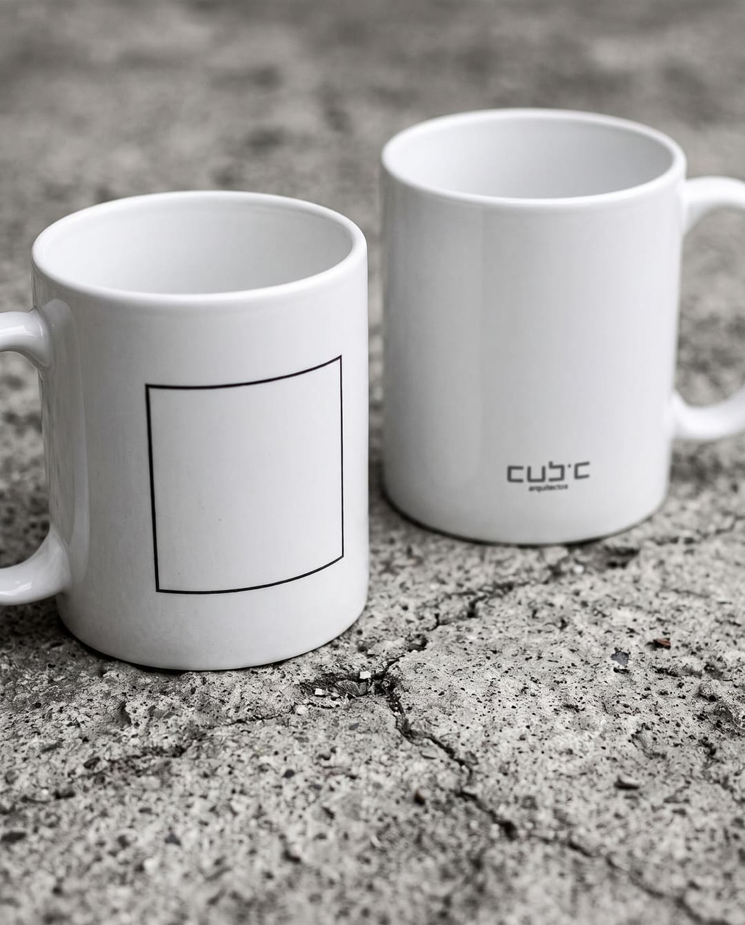 Duas canecas brancas sobre betão: uma com um quadrado desenhado a traço fino, a outra com o logótipo da Cubic.
