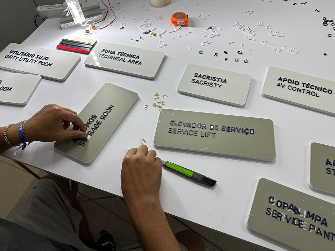 Mãos a colar letras em placas bilingues: Elevador de serviço, Sacristia, Zona técnica, Apoio técnico.