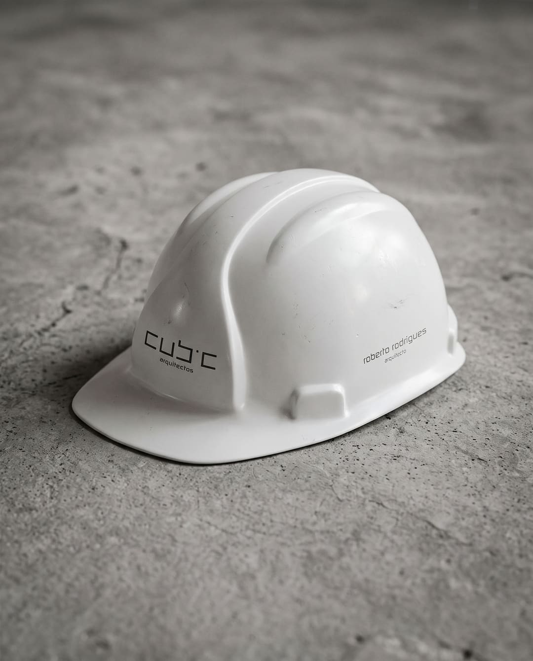 Um capacete de obra branco sobre betão, com o logótipo da Cubic num lado e «roberto rodrigues arquitecto» no outro.