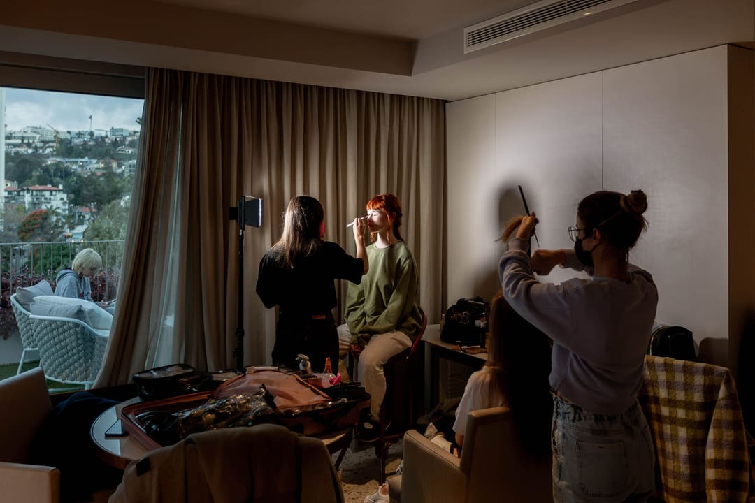 Num quarto com vista para a cidade, uma maquilhadora trabalha a cara de uma modelo ruiva sob um painel de luz, enquanto, à direita, outra pessoa penteia o cabelo de alguém sentado.