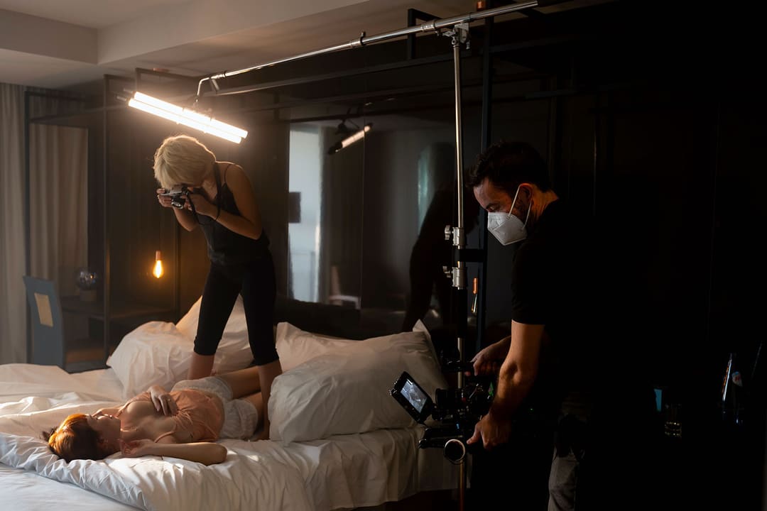 Uma modelo ruiva deitada numa cama de lençóis brancos, fotografada de cima por uma fotógrafa de pé na cama, sob uma barra de luz, e filmada por um operador de câmara à direita.